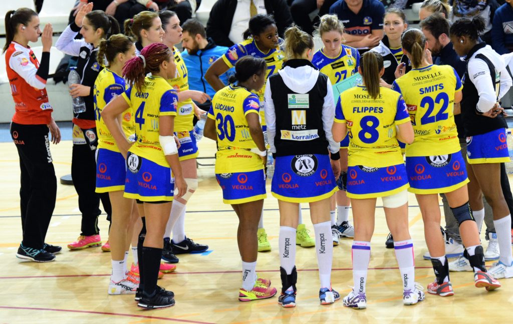 &Eacute;quipe f&eacute;minine du Metz Handball en temps mort, joueuses en maillot jaune et bleu avec jupette, rassembl&eacute;es autour de l&rsquo;entra&icirc;neur sur le terrain.
